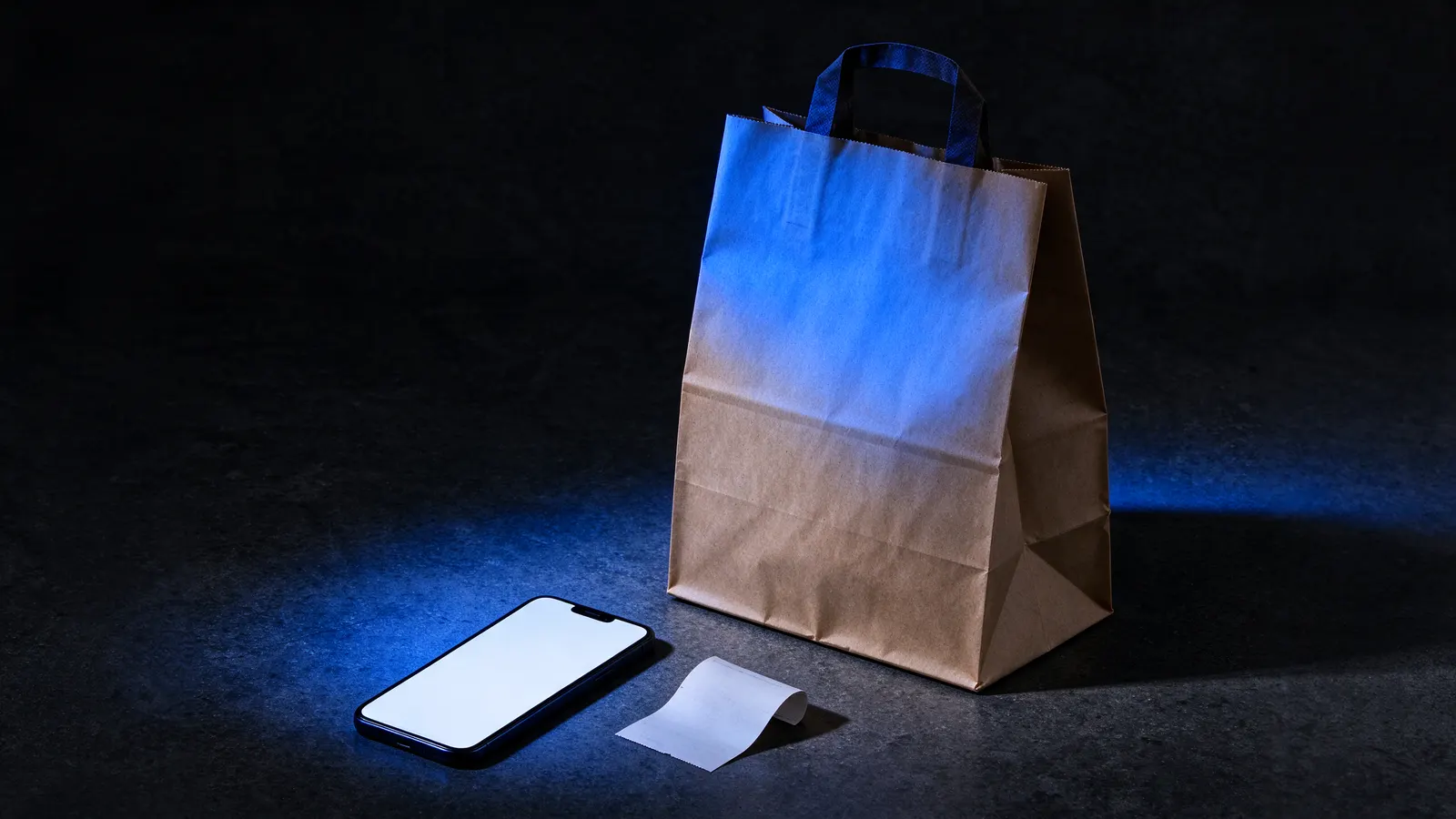 Brown paper delivery bag on a dark stone counter, next to a phone with its screen on and a paper coupon, under blue side light