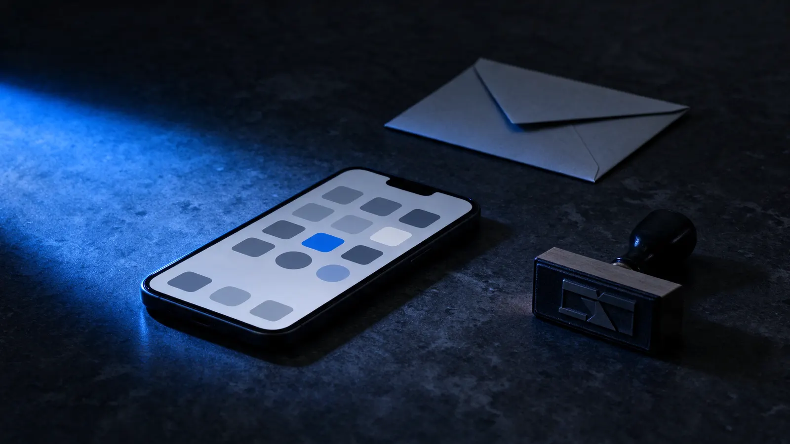Celular com a tela acesa mostrando uma grade de ícones cinza e um azul, sobre uma mesa de pedra escura, ao lado de um envelope e de um carimbo, com uma faixa de luz azul vindo da esquerda