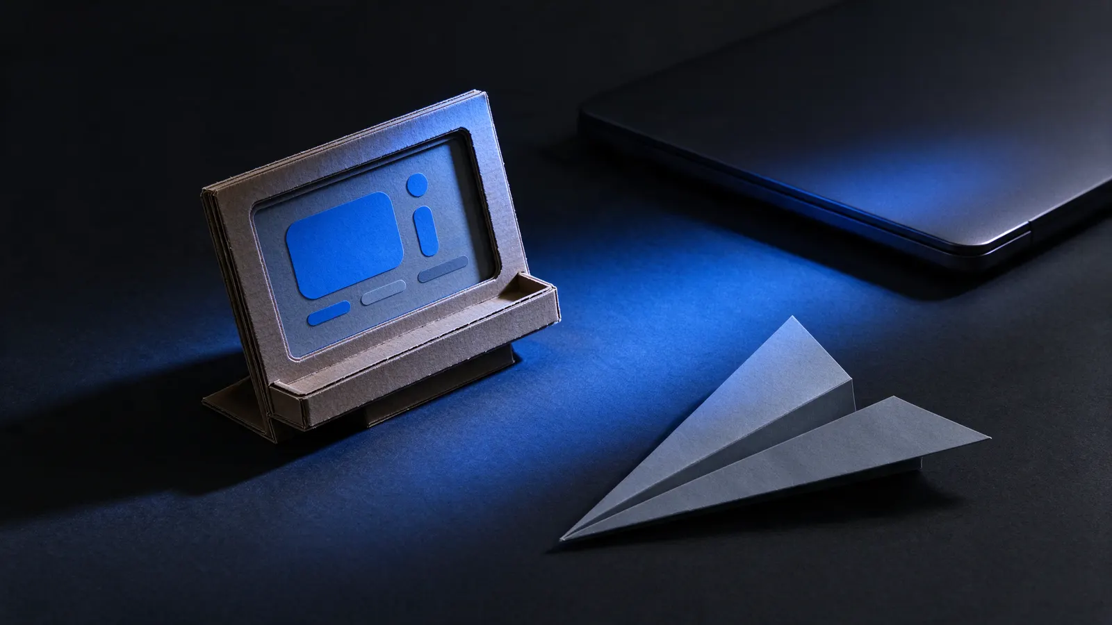 Protótipo de tela feito de papelão, com blocos azuis no lugar dos botões, ao lado de um avião de papel e de um notebook fechado sobre uma mesa escura iluminada por luz azul