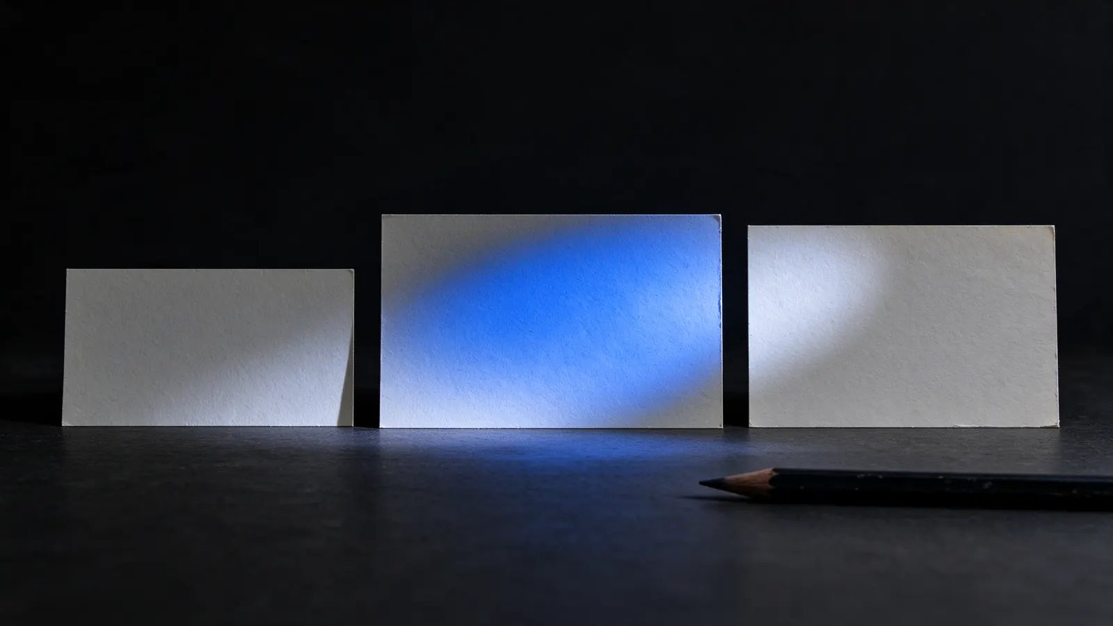Três cartões em branco de tamanhos diferentes em pé sobre uma mesa escura, o do meio iluminado por luz azul, com um lápis à frente
