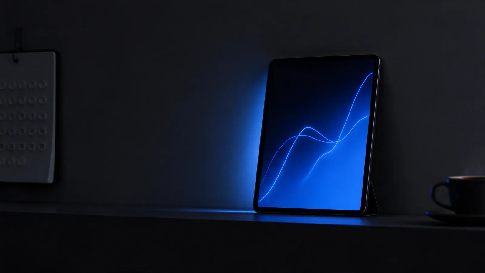 Tablet apoiado numa prateleira escura com um gráfico de linhas azul aceso na tela, ao lado de um calendário de parede e uma xícara de café